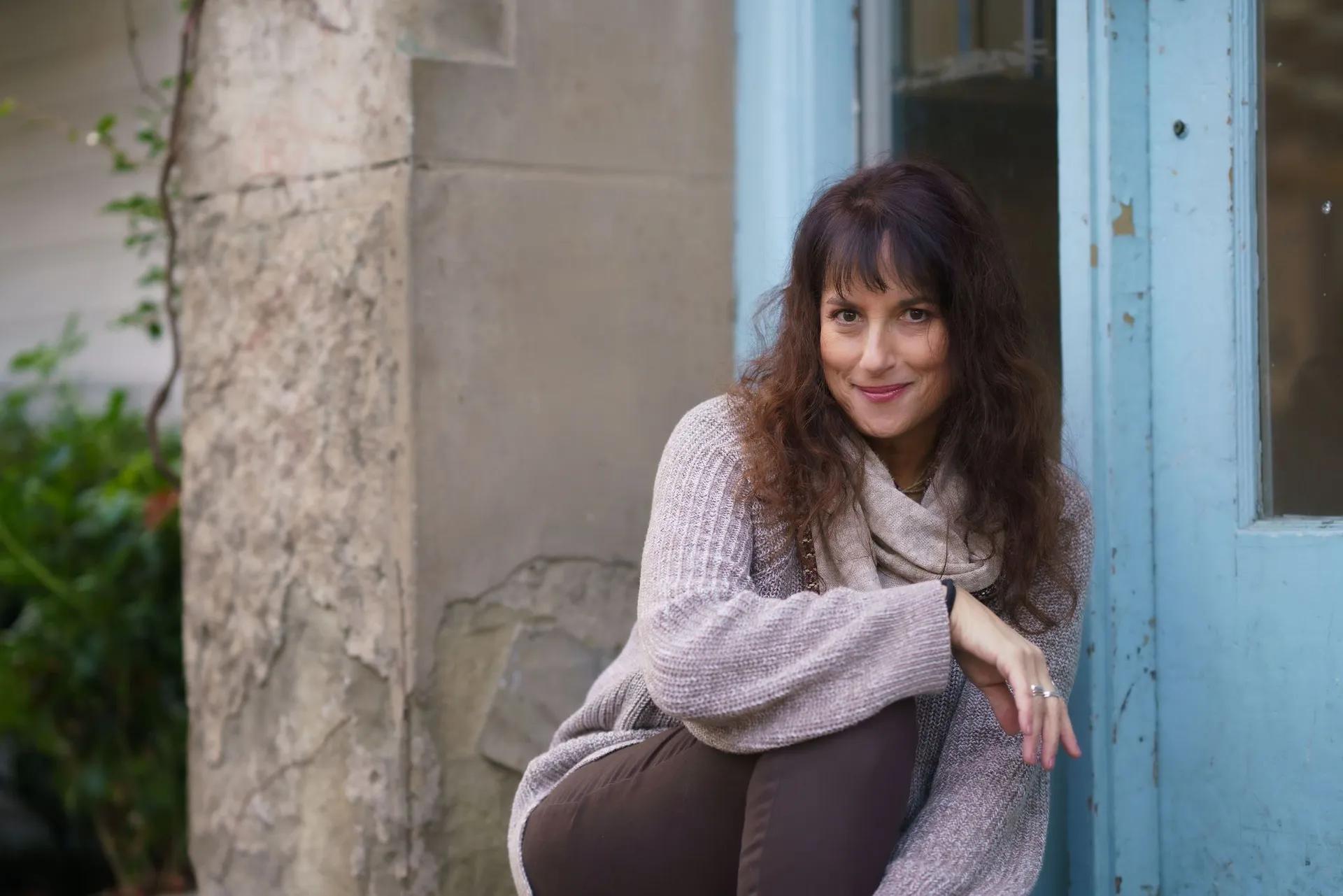 Issy Kleiman, MA, LMFT, AAMFT Approved Supervisor, seated beside a pale blue door at the practice, wearing a camel cardigan and scarf